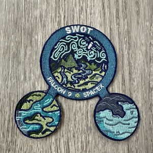 May include: Embroidered patch with a blue and green design. The patch features the text "SWOT" at the top, with a landscape scene inside a circle. The words "FALCON 9" and "SPACEX" are also visible.