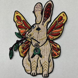 Könnte beinhalten: Gestickter Aufnäher mit einem Hasen mit Schmetterlingsflügeln, der einen Zweig hält. Der Hase ist beige mit goldenen und schwarzen Akzenten, blauen Augen und einem grünen Anhänger. Die Flügel sind rot, orange und gelb.