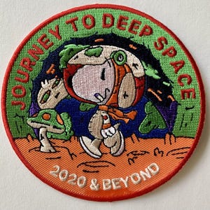 Op de afbeelding: Geborduurde ronde patch met een stripfiguur in een astronautenpak, omringd door een groene en blauwe ruimtescène. De patch heeft de tekst "JOURNEY TO DEEP SPACE 2020 & BEYOND" in een cirkelvormig ontwerp, met een rode rand en een oranje achtergrond.