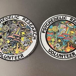 Op de afbeelding: Twee ronde geborduurde patches met de tekst "PSYCHEDELIC RESEARCH VOLUNTEER". De patches tonen kleurrijke, ingewikkelde ontwerpen met paddenstoelen, aliens en psychedelische beelden. Eén patch heeft een zwarte achtergrond, de andere een witte achtergrond.