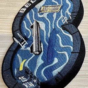 Könnte beinhalten: Gestickter Aufnäher mit dem Text "BELLY FLOP" in Weiß auf schwarzem Rand. Der Aufnäher zeigt ein blau-weißes Design mit einer Rakete und anderen Elementen.