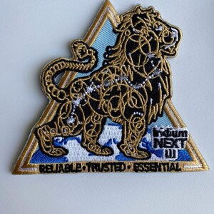 May include: A gold embroidered patch with a lion design on a blue background. The patch is triangular and has the text "Trivium Next III" and "Reliable. Trusted. Essential" on it.