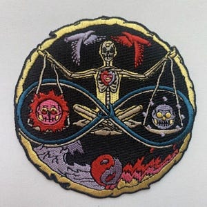 Op de afbeelding: Een ronde geborduurde patch met een skeletfiguur die een weegschaal vasthoudt met een zon en een maan aan elke kant. Het skelet wordt omringd door een oneindigheidssymbool en een yin-yang-symbool. De patch heeft een gouden rand en bevindt zich op een witte achtergrond.