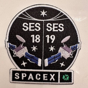 Puede incluir: Un parche bordado en blanco y negro con el texto "SES 18 SES 19" y "SpaceX" con un trébol verde. El parche presenta dos antenas parabólicas estilizadas.