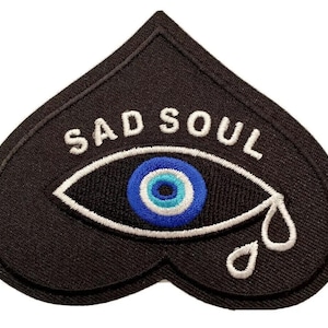 May include: Black heart-shaped patch with the words "SAD SOUL" embroidered in white. The patch features an eye design with a blue and white iris, and two white teardrops. The patch is likely for clothing or accessories.