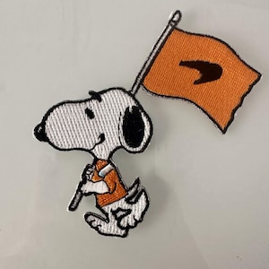 May include: An embroidered patch featuring Snoopy, a cartoon beagle, holding a flag with an orange background and a black swoosh logo. Snoopy is wearing an orange shirt and white pants.