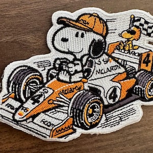 May include: Embroidered patch featuring Snoopy driving a race car with the word "McLaren" and the number "4". Snoopy wears an orange cap and another Snoopy waves a checkered flag. The car is white and orange.