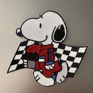 Puede incluir: Parche bordado con Snoopy con un traje de carreras rojo, sosteniendo una bandera a cuadros. El parche tiene una combinación de colores blanco, negro, rojo y azul. El personaje de Snoopy está sonriendo y parece estar corriendo.