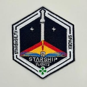 Könnte beinhalten: Ein schwarz-weißes Abzeichen mit einem weißen sechseckigen Rand. Die Mitte des Abzeichens zeigt eine weiße Rakete mit einer rot-gelb-blauen Flamme. Der Text "STARSHIP FLIGHT 2" ist in Weiß auf einem gelben Banner gedruckt. Der Text "STARBASE, TX" ist in Weiß auf der linken Seite des Randes gedruckt und "SPACEX" ist in Weiß auf der rechten Seite des Randes gedruckt. Ein grünes vierblättriges Kleeblatt ist in der Mitte unten des Abzeichens gedruckt.