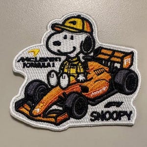 May include: Embroidered patch featuring Snoopy in a racing suit and helmet, driving an orange McLaren Formula 1 car. The patch includes the text "McLaren Formula 1" and "Snoopy" in white lettering. The design is outlined in white.