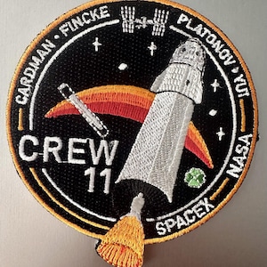 Puede incluir: Parche bordado con una nave espacial y el texto "CREW 11", con los nombres Cardman, Fincke, Platonov y Yui. El parche incluye los logotipos de la NASA y SpaceX, sobre un fondo negro con un borde dorado.