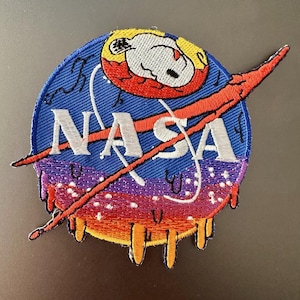May include: Embroidered patch featuring the NASA logo with a Snoopy character in a space helmet. The patch has a blue background with white text, a red and orange swoosh, and a melting effect with purple, orange, and yellow colors.