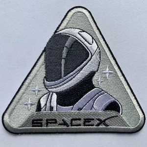 May include: A silver and gray triangular patch with an embroidered image of an astronaut wearing a helmet. The astronaut is facing the left side of the patch. The text "SPACEX" is embroidered in black at the bottom of the patch.