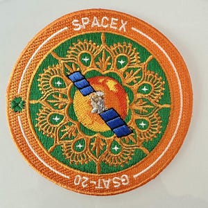 Peut inclure: Écusson brodé circulaire avec le mot "SPACEX" en haut et "GSAT-20" en bas. Le motif central comprend un satellite, un élément en forme de soleil et des motifs complexes verts et dorés, le tout sur un fond orange. Un objet décoratif.