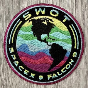 May include: Circular embroidered patch with a colourful Earth design. The patch features the text "SWOT SPACEX 9 FALCONS" in a circular pattern. The Earth graphic is set against a black background with a pink border.