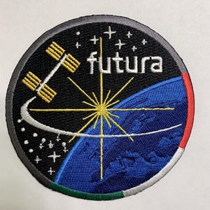 Puede incluir: Un parche bordado negro y azul con un estallido de estrella blanco, una estación espacial amarilla y el texto "futura" en blanco. El parche tiene un borde rojo, verde y blanco.