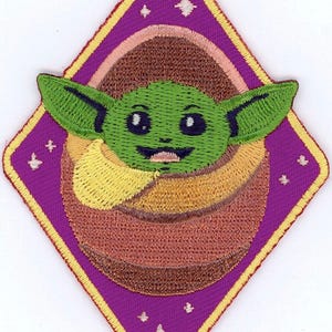 May include: Embroidered patch of a green alien character with large ears and big eyes, set within a purple diamond shape. The design includes a brown and yellow background, with stars and a yellow border.