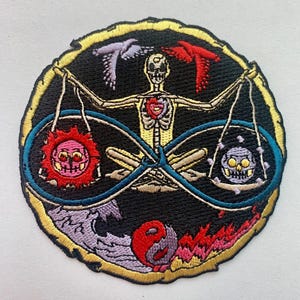 Könnte beinhalten: Gestickter Aufnäher mit einer Skelettfigur, die Waagen hält, mit einer Sonne und einem Totenkopf auf jeder Seite eines Unendlichkeitszeichens. Der Aufnäher hat einen schwarzen Hintergrund mit einem goldenen Rand und enthält ein Yin-Yang-Symbol und Flammen.