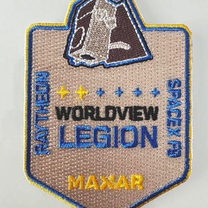 Puede incluir: Un parche bordado de color beige con un borde azul y amarillo. El parche presenta un contorno marrón de un satélite con el texto "WORLDVIEW LEGION" y "MAXAR" debajo. El texto "RAY THEON" está en el lado izquierdo del parche y "SPACEX" en el lado derecho.