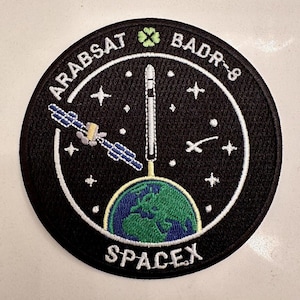 Puede incluir: Un parche bordado en blanco y negro con un círculo blanco que contiene un globo terráqueo verde, un cohete blanco, un satélite blanco y estrellas blancas. El texto "ARABSAT BADIR-8 SPACEX" está escrito en blanco alrededor del círculo. Un trébol de cuatro hojas verde está por encima del texto.