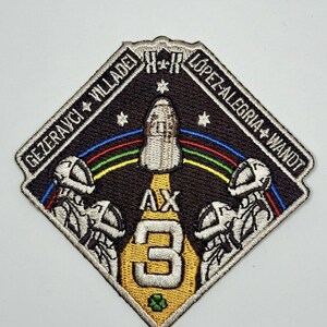 Peut inclure: Un écusson en forme de diamant avec un fond noir et un vaisseau spatial blanc au centre. Le vaisseau spatial est entouré d'un arc-en-ciel et d'étoiles. Le texte "AX-3" est au centre de l'écusson, avec les noms des astronautes "GEZERAUCH + VILLADEI + LOPEZ-ALEGRIA + WANDT" autour du bord.