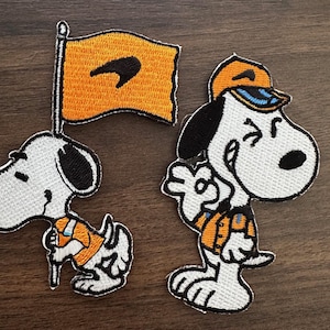 May include: Two embroidered Snoopy patches. One shows Snoopy holding a flag with a black design on an orange background. The other patch depicts Snoopy wearing a cap and waving. Both patches have black, white, and orange details.