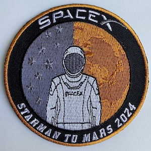 May include: A round, black and gold embroidered patch with a white astronaut in the center. The patch says "SpaceX Starman to Mars 2024" around the edge.