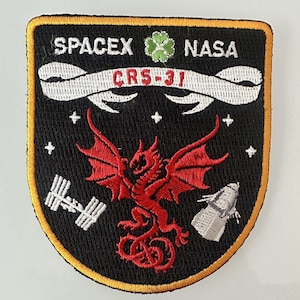 Può includere: Toppa ricamata con i loghi SpaceX e NASA, e la scritta "CRS-31". Un drago rosso è al centro, con una stazione spaziale e un veicolo spaziale su entrambi i lati. La toppa ha uno sfondo nero e un bordo dorato.