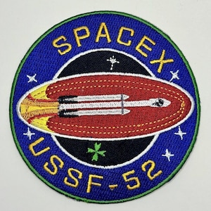 Puede incluir: Un parche bordado azul y blanco con el texto "SpaceX USSF-52" y una nave espacial roja y amarilla en el centro.