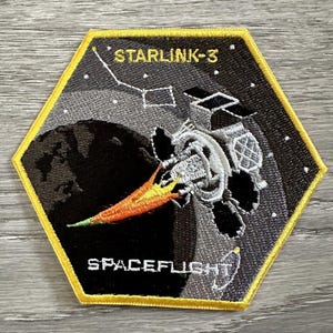 May include: A hexagonal patch with a yellow border, depicting a spacecraft with a trail of fire, against a backdrop of a gray and white Earth. The text "STARLINK-3" and "SPACEFLIGHT" are printed on the patch.