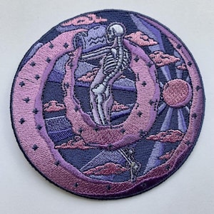 Puede incluir: Parche bordado con una figura de esqueleto sosteniendo una luna creciente en un esquema de colores morado y azul. La luna está rodeada de nubes y estrellas.