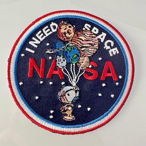 May include: A blue and white embroidered patch with the text "I NEED SPACE NASA" in red and white. A cartoon astronaut is holding a bunch of planets with strings.