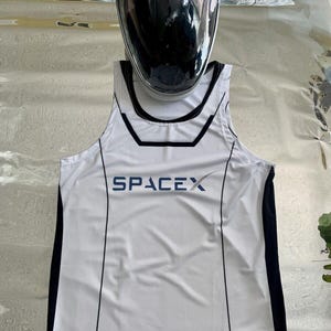 May include: A white SpaceX tank top with black trim, the SpaceX logo, and an American flag patch. A white astronaut helmet with a dark visor rests above the tank top. A NASA patch is also visible.