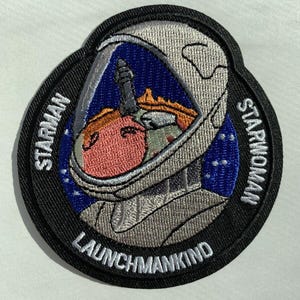 May include: A black and white embroidered patch with a cartoon astronaut wearing a helmet. The patch has the text "Starman Launchmankind Starwoman" around the edge.