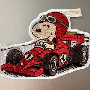 May include: Embroidered patch featuring Snoopy in a red race car with the number 1. Snoopy wears a red helmet and goggles. The car has black tires and a white outline.