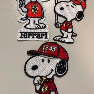 Puede incluir: Parches bordados con Snoopy con atuendos Ferrari rojos. Uno muestra a Snoopy con un signo de la paz y la palabra "Ferrari". Otro sostiene una bandera de Ferrari, y el tercero lleva una gorra de Ferrari.