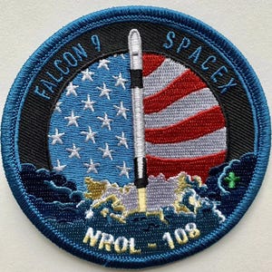 Pode incluir: Um remendo bordado azul e preto com um foguete branco lançando através de uma bandeira americana estilizada. O texto "FALCON 9 SPACEX" é escrito ao redor da parte superior do remendo. O texto "NRL 108" é escrito na parte inferior do remendo.