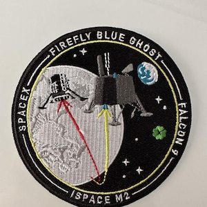May include: A black and white embroidered patch featuring a white moon, a rocket, and a smaller blue planet. The text "FIREFLY BLUE GHOST" is in a yellow circle around the top of the patch. The text "SPACE M2" is in a yellow circle around the bottom of the patch. The text "SPACEX FALCON 9" is in a yellow circle around the right side of the patch.