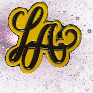 May include: A yellow and black enamel pin featuring the letters "LA" in a cursive script. The pin has a glossy finish and a black outline, set against a speckled, light purple and white background.