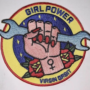 Puede incluir: Un parche bordado amarillo y rojo con un puño que sostiene una llave inglesa. El parche dice "Girl Power" y "Virgin Orbit" en un círculo.