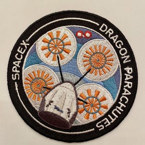 May include: A black and white embroidered patch with a white capsule and four orange and white parachutes. The text "SPACEX" is written in white on a black background and "DRAGON PARACHUTES" is written in white on a black background.