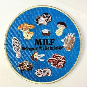 May include: A round, embroidered patch with a blue background and a gold border. The patch features various mushroom illustrations and the text "MILF Mushrooms I'd Like to Forage."