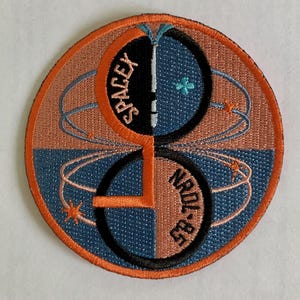 Puede incluir: Un parche redondo con borde naranja y un diseño en blanco y negro. El diseño presenta dos círculos superpuestos con una línea blanca y una estrella azul en el centro. El texto "SPACEX" está escrito verticalmente en el círculo superior y "NRL-825" está escrito verticalmente en el círculo inferior.