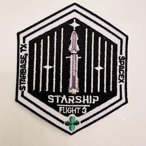 Puede incluir: Un parche bordado blanco y negro con una nave espacial blanca en el centro. El parche tiene forma de hexágono y tiene el texto "Starship Flight 3" y "SpaceX".