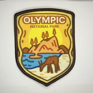Könnte beinhalten: Gestickter Aufnäher in Schildform mit Darstellung des Olympic National Park. Das Design umfasst eine Bergkette, einen See, ein Reh und den Text "OLYMPIC NATIONAL PARK". Der Aufnäher hat einen braunen Rand. Die Farbpalette umfasst Gelb, Orange, Blau und Braun.