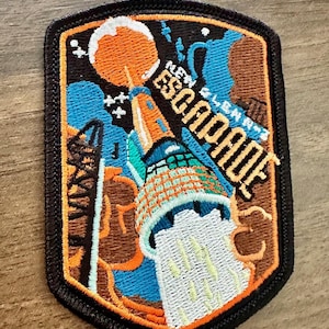 Puede incluir: Parche bordado con forma de escudo con el texto "New Glenn's Escapade". El parche presenta un cohete, un planeta y elementos espaciales en naranja, azul y marrón, con un borde negro. El diseño es detallado y colorido.