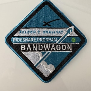 May include: A blue and black diamond-shaped patch with white embroidery. The patch features a rocket ship with a white plume, a white cloud, and the text "Falcon 9 SmallSat Rideshare Program Bandwagon".