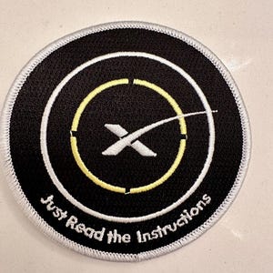May include: A black and white embroidered patch with a white X inside a yellow circle and the text "Just Read the Instructions" around the outside.