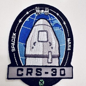 May include: A black and white embroidered patch with a white spaceship in front of a blue and white globe. The text "SPACEX" is on the left side of the patch and "NASA" is on the right side. The text "CRS-30" is in a gray banner at the bottom of the patch.