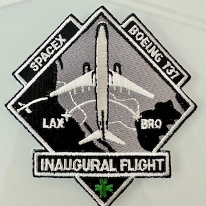 Op de afbeelding: Een witte en grijze ruitvormige patch met de tekst "SPACEX" en "BOEING 737" op de bovenste hoeken. De patch toont een wit vliegtuig met een grijze kaart van de Verenigde Staten op de achtergrond. De tekst "INAUGURAL FLIGHT" staat onderaan de patch. Een groen medisch symbool bevindt zich in het midden van de patch.